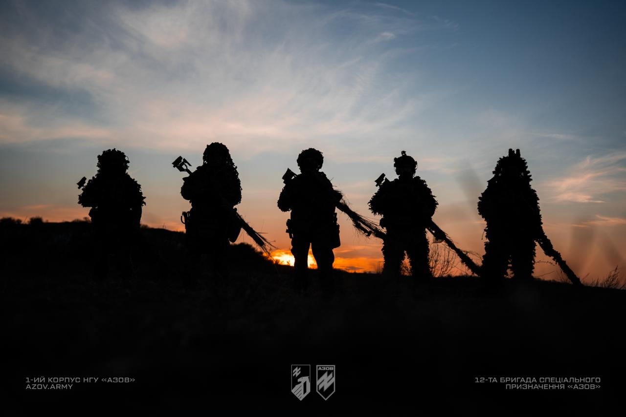  Силуети пʼятьох бійців, що стоять зі зброєю у променях заходу сонця